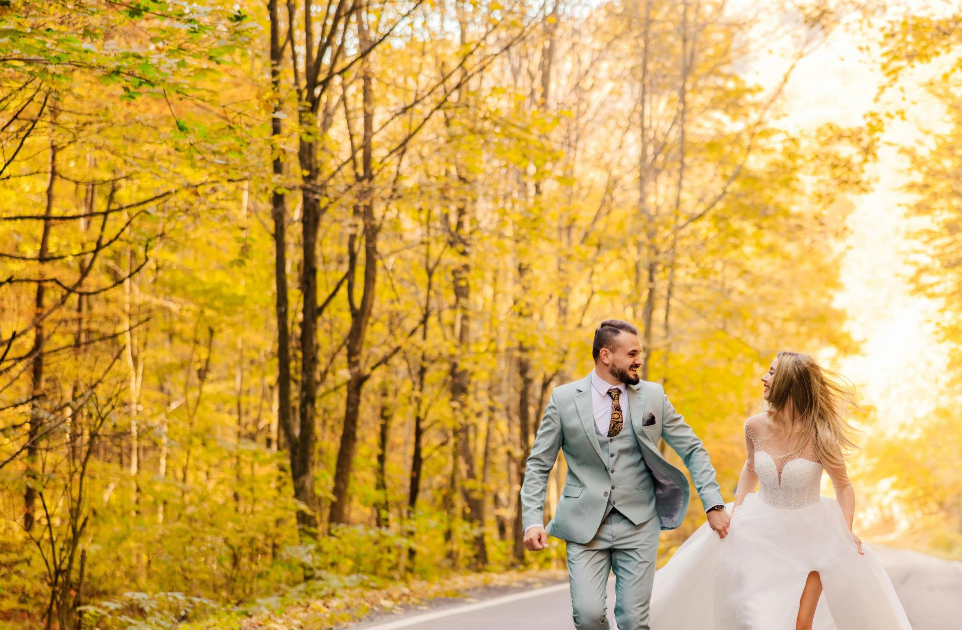 A joyful bride and groom run down a scenic road in a colorful autumn forest, capturing their wedding day joy.